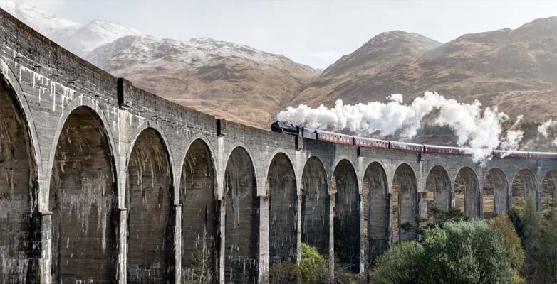 A steam train crosses a viaduct in open countryside, with plumes of steam following in its wake