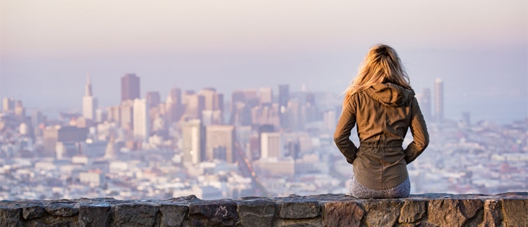 Image of a lady sitting on a wall with her hands in her pocket facing away and looking at a city skyline