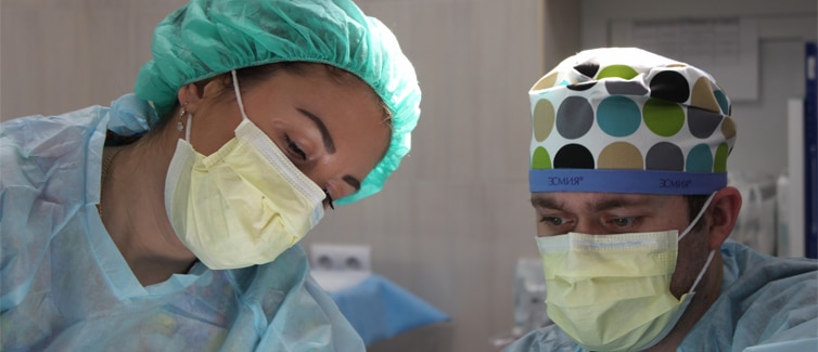 Two people wearing medical face masks, scrub caps and surgical gowns, looking downwards