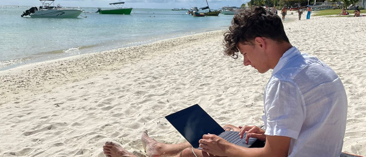 A student sat on a white sandy beach looking at their laptop which is resting on their lap