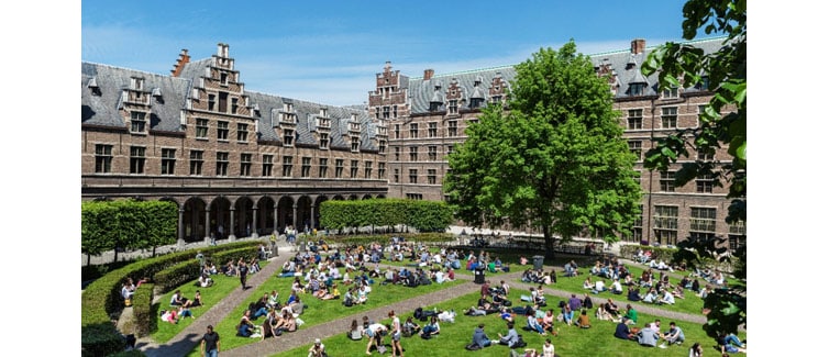 Image of a large, intricately designed building with a grass courtyard in front, with lots of people sat down in groups on the grass