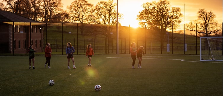 Image of 6 footballers standing in a line on a football pitch, with footballs in the foreground and a sunset in the background