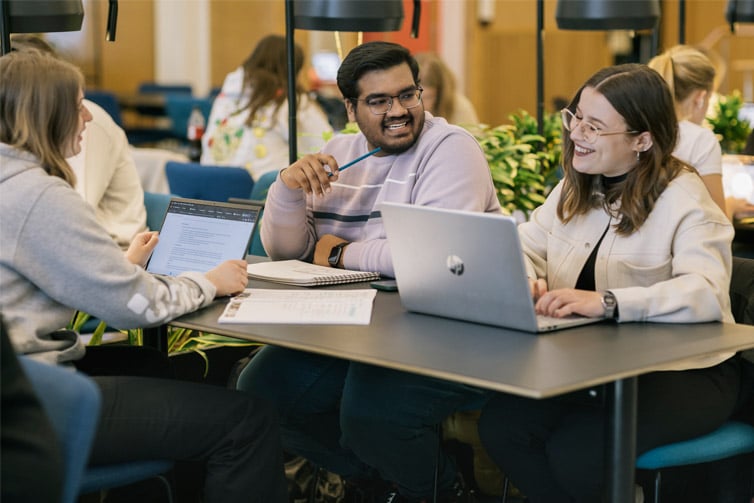 Image of three students sat at a table together with laptops open smiling at each other