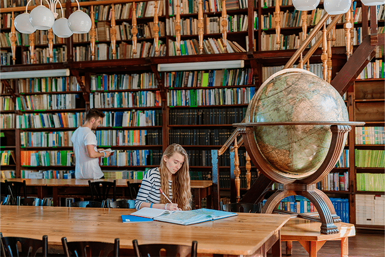 Image of a student sat at a wooden table in a library writing in a notebook