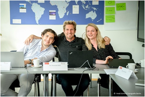 Image of three students sat at a table leaning in towards each other and smiling at the camera