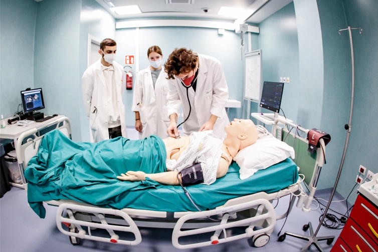 Image of 3 students wearing white medical coats and masks in a mock hospital room with a hospital bed and body dummy